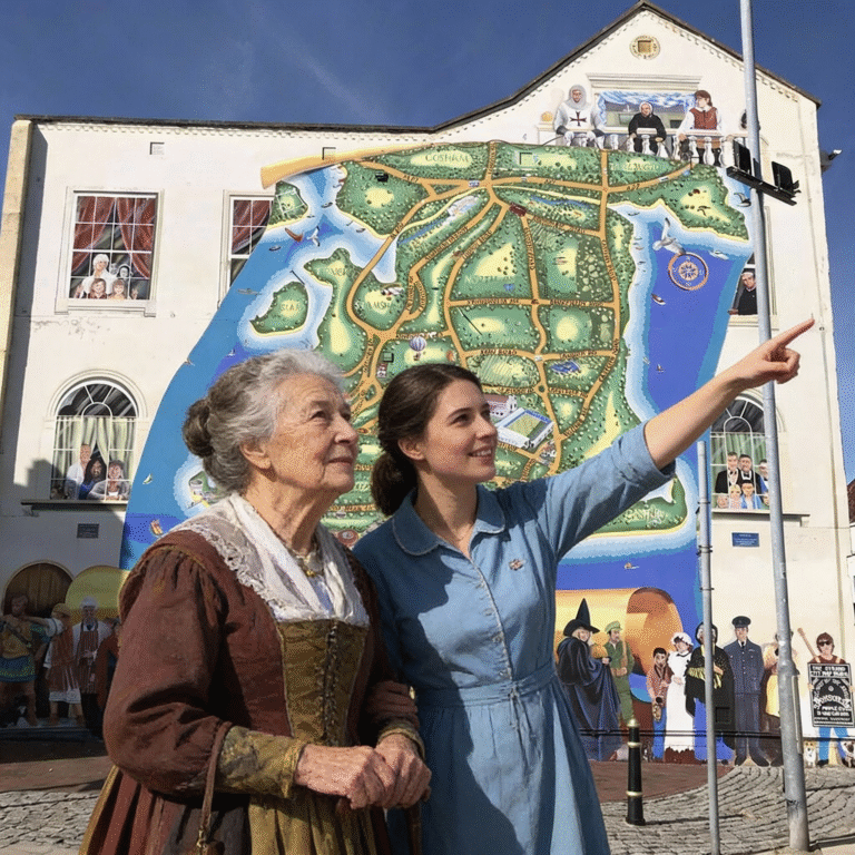 elderly lady in period dress standing with a companion who is pointing into the distance, next to a large building mural